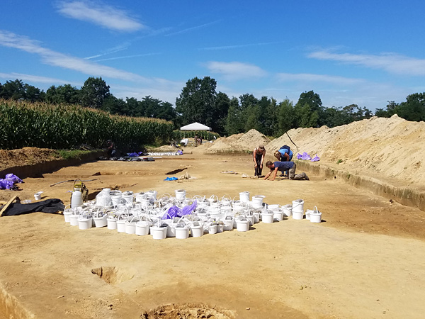 Several people work in a large, shallow, rectangular pit surrounded by piles of earth. In the middle are many white buckets